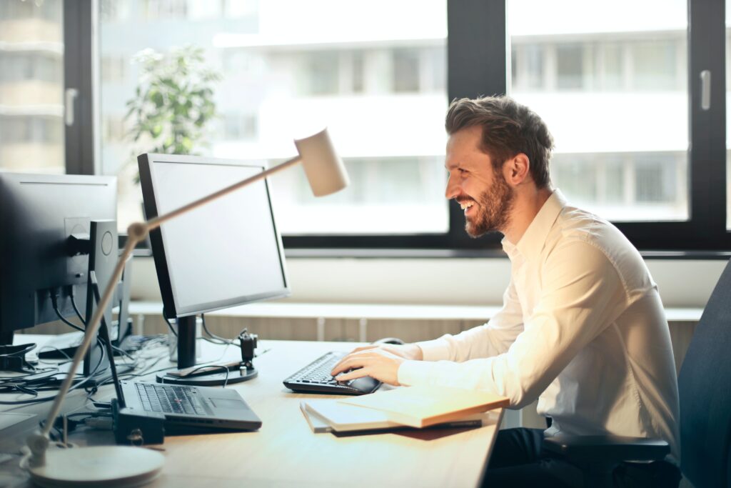 SOLUTIONS METRIX photo, Man in White Dress Shirt Sitting on Black Rolling Chair While Facing Black Computer Set and Smiling